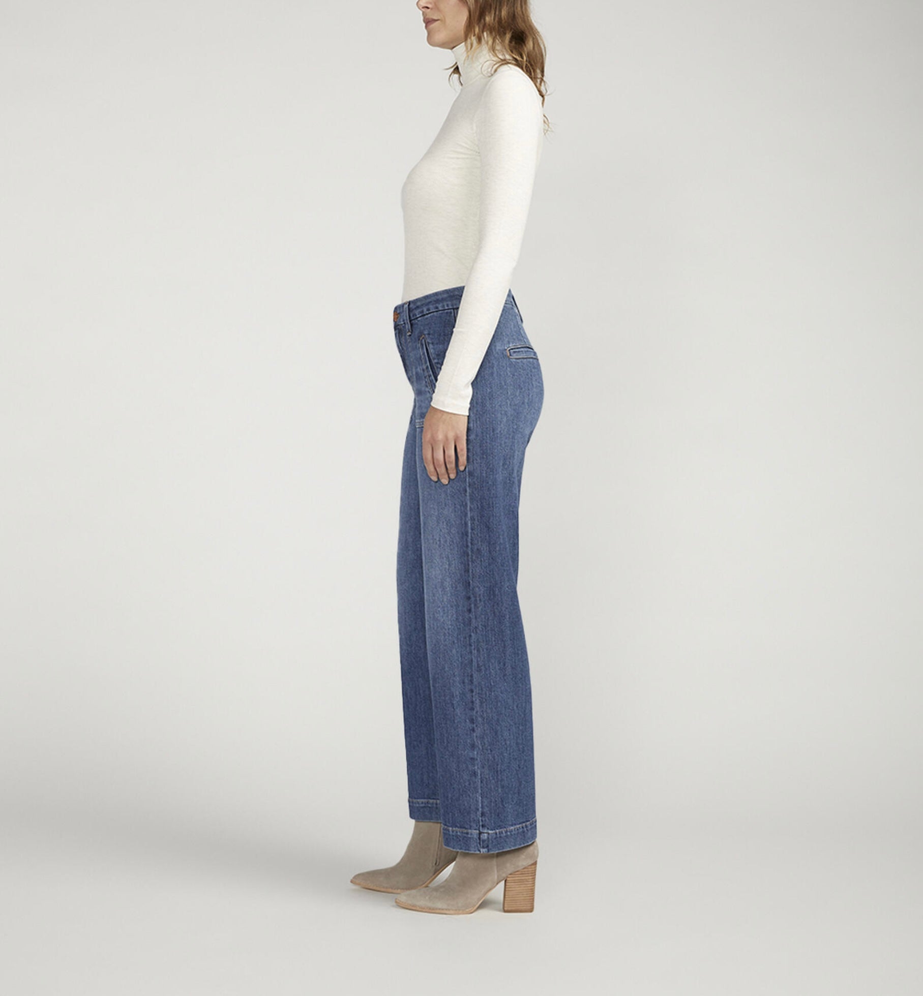 A person standing against a plain background is wearing a white turtleneck long-sleeve shirt, **JAG's Sophia High Rise Wide Leg Jeans in medium indigo wash with front pockets**, and light-colored ankle boots. Their hands are in their pockets and they are facing forward.