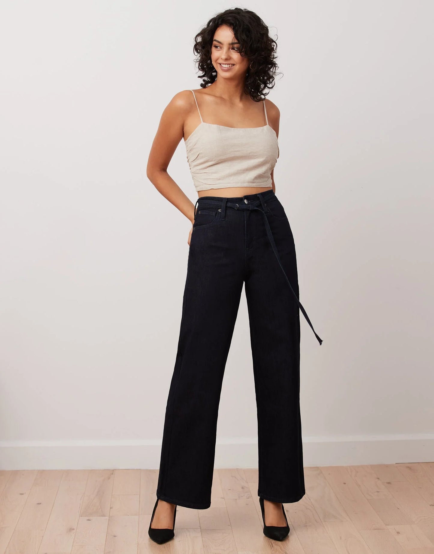 A woman stands smiling in a beige spaghetti strap top and high-rise Lily Wide Leg Jeans in Abel Navy by Yoga Jeans, styled with a belt and black heels, on a light wood floor against a plain white wall.