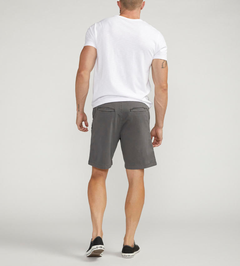 A man is standing while wearing a white short-sleeve t-shirt, Silver Jeans Co.'s PULL-ON CHINO SHORT DARK GREY Style M52155CBT639, and black sneakers with white laces. He has a tattoo on his right arm and a beard. His left hand is in his pocket, and he is looking to his left. The background is plain and light-colored.