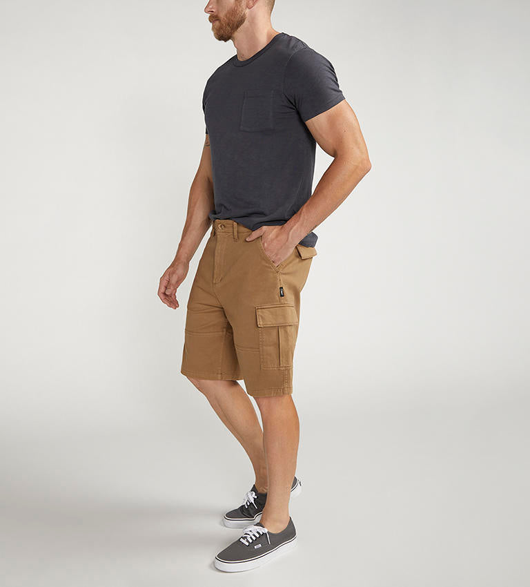 A man stands against a plain background wearing a dark gray t-shirt with a chest pocket, relaxed fit CARGO SHORT DESERT styled by Silver Jeans Co. (product code: M53151CBT639), and black sneakers with white soles. He appears to be adjusting his shirt with his right hand.