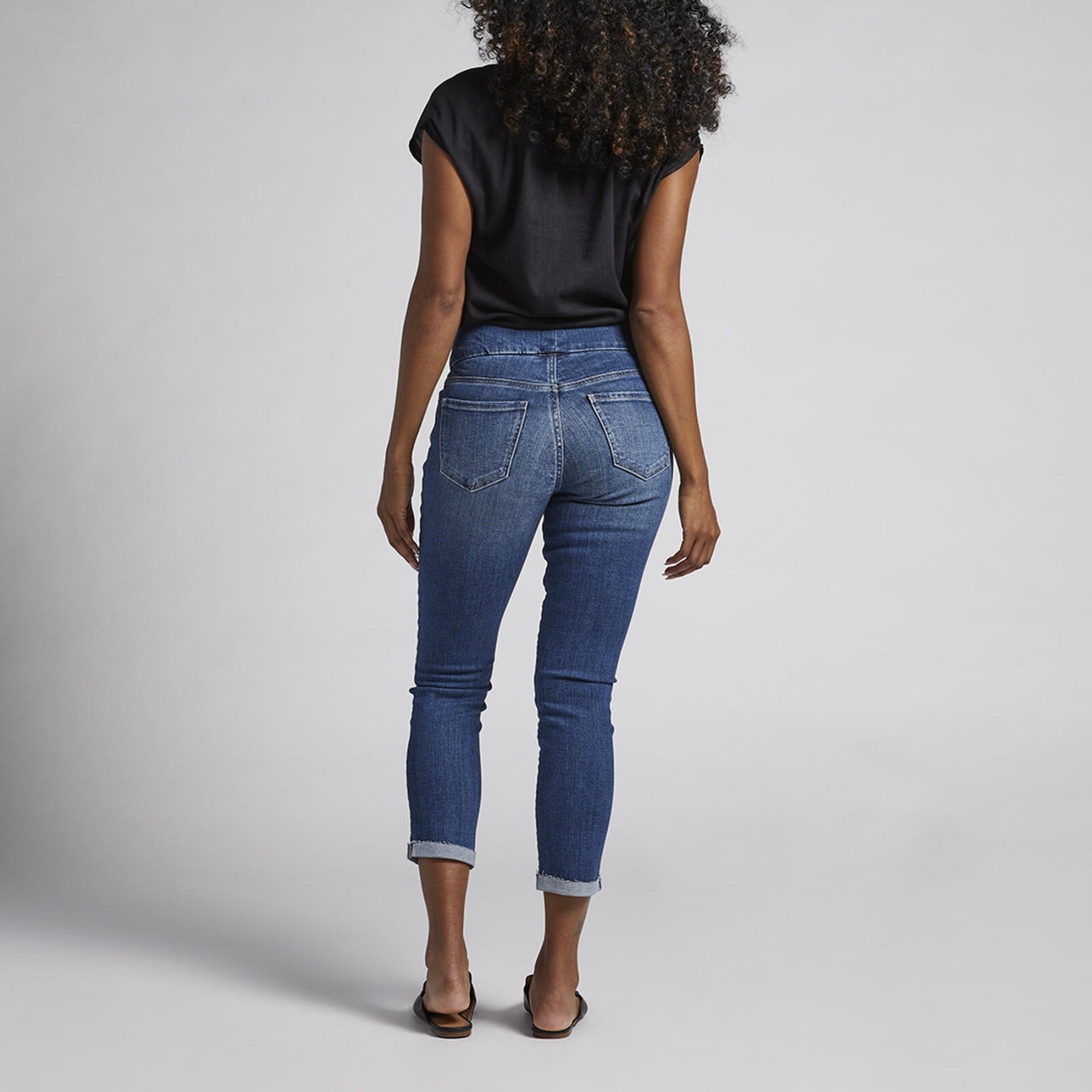 A person with curly hair is wearing a black short-sleeve blouse, Amelia Mid Rise Slim Ankle Pull-On Jeans by JAG, and black loafers. They are standing against a plain light grey background, smiling, with their hands relaxed by their sides.