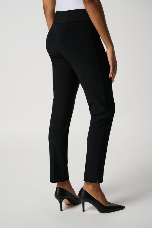 A person is standing and posing in Joseph Ribkoff's Classic Tailored Slim Pant (144092NOS-6), made from flattering silky knit fabric, paired with black high-heeled shoes. They are wearing a white top. The background is a plain light color.