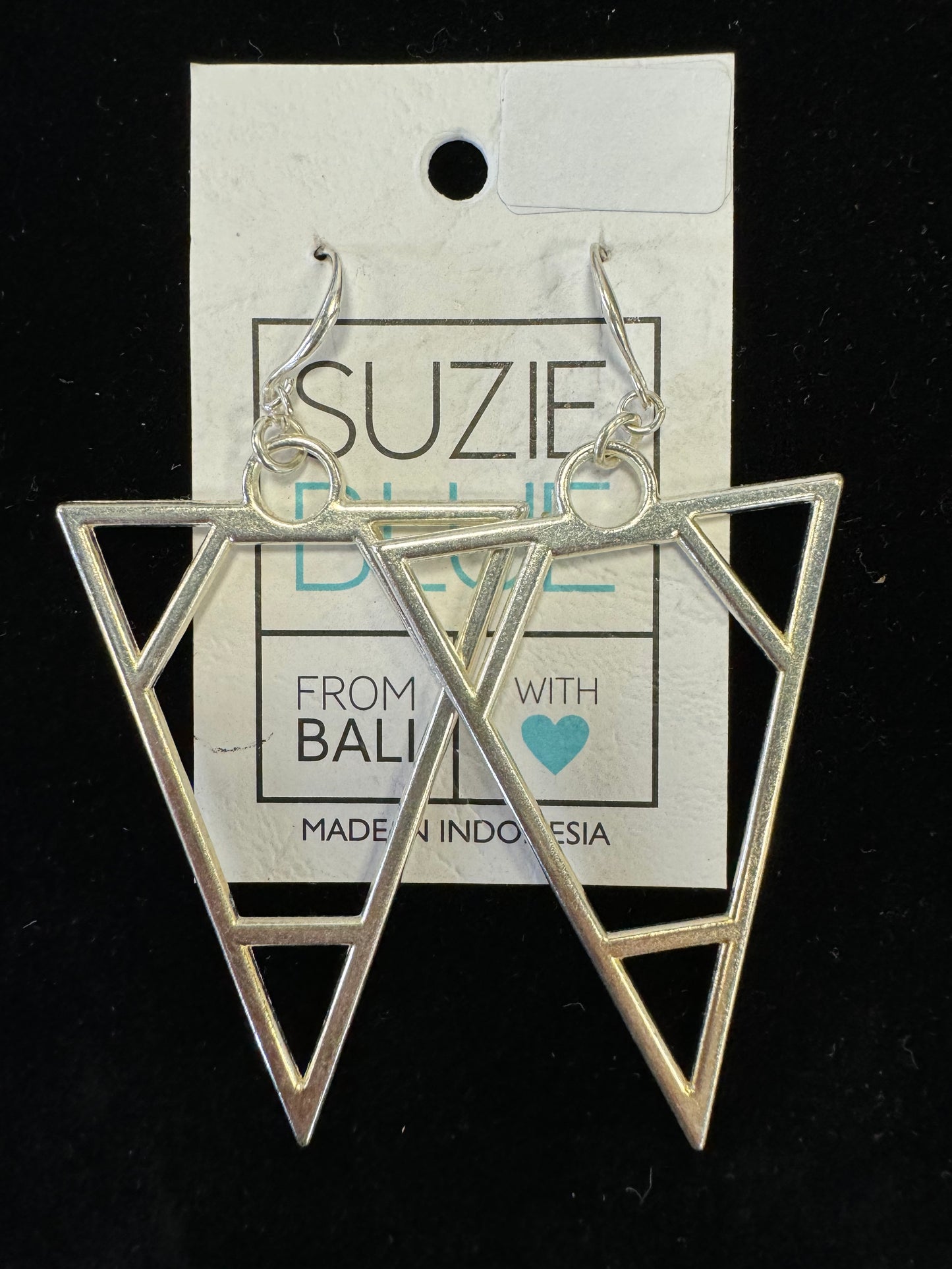 A pair of Turned Triangle Dangling Earrings from Suzie Blue are displayed on a black background. They hang from a card labeled "Suzie Blue" with text indicating "From Bali with Love" and "Made in Indonesia." The card features a heart in teal blue.