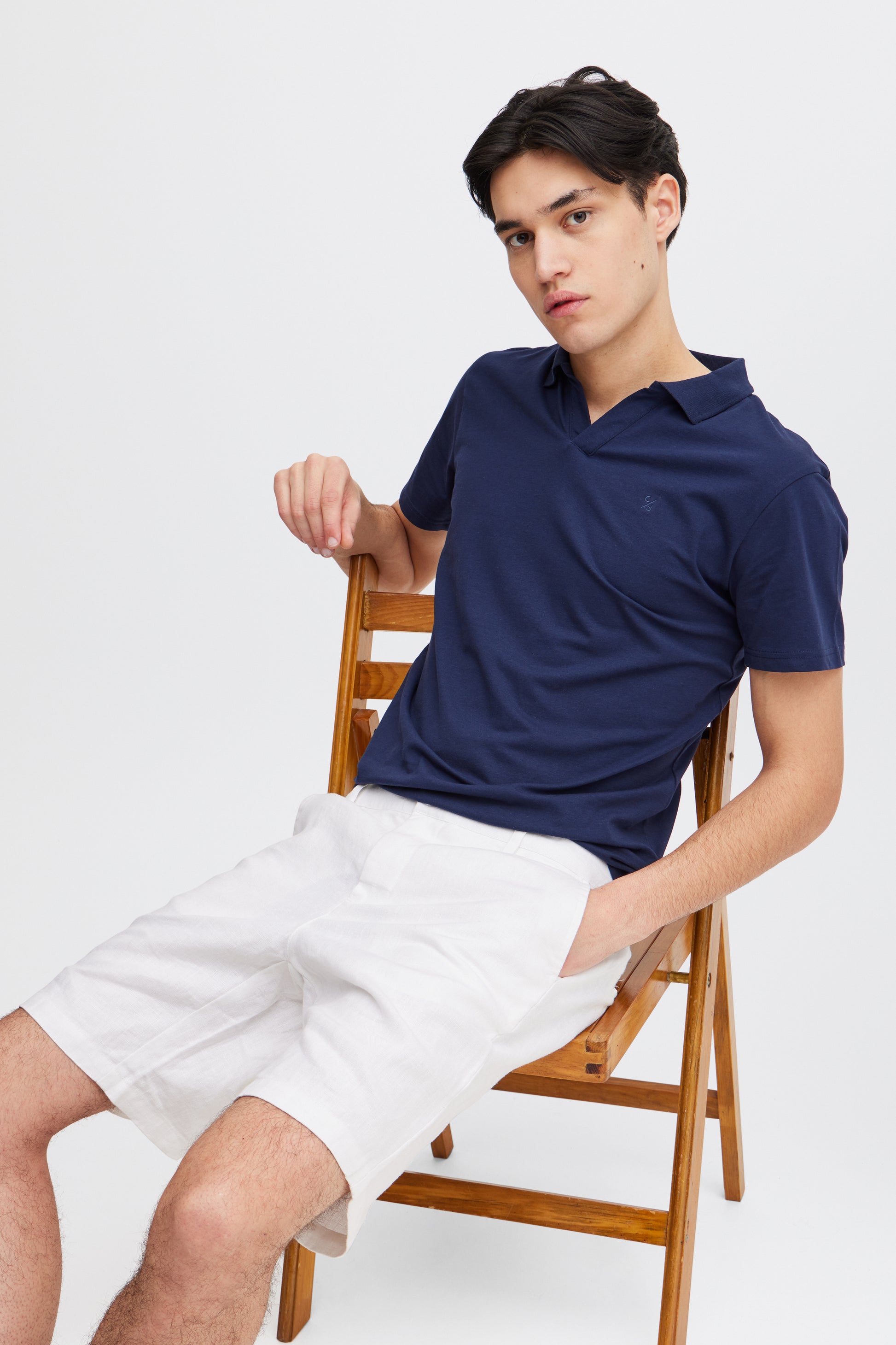 A young man with short dark hair sits on a wooden chair wearing white Linen Shorts 20505166 by Casual Fridays and a navy polo, hand in pocket, looking relaxed—an effortless style perfect for casual Friday comfort.