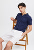 A young man with short dark hair sits on a wooden chair wearing white Linen Shorts 20505166 by Casual Fridays and a navy polo, hand in pocket, looking relaxed—an effortless style perfect for casual Friday comfort.