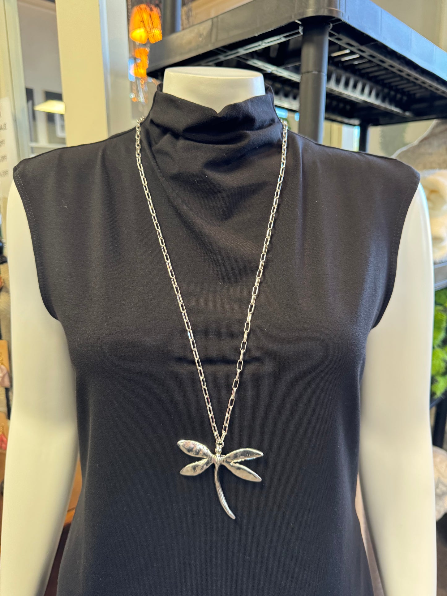 A headless mannequin is showcasing a sleeveless black turtleneck dress, enhanced by the striking Andy 13Necklace from Elise Accessories, featuring a dragonfly pendant. This captivating piece from our latest fashion jewelry collection stands out against the shelves lined with various items behind it.