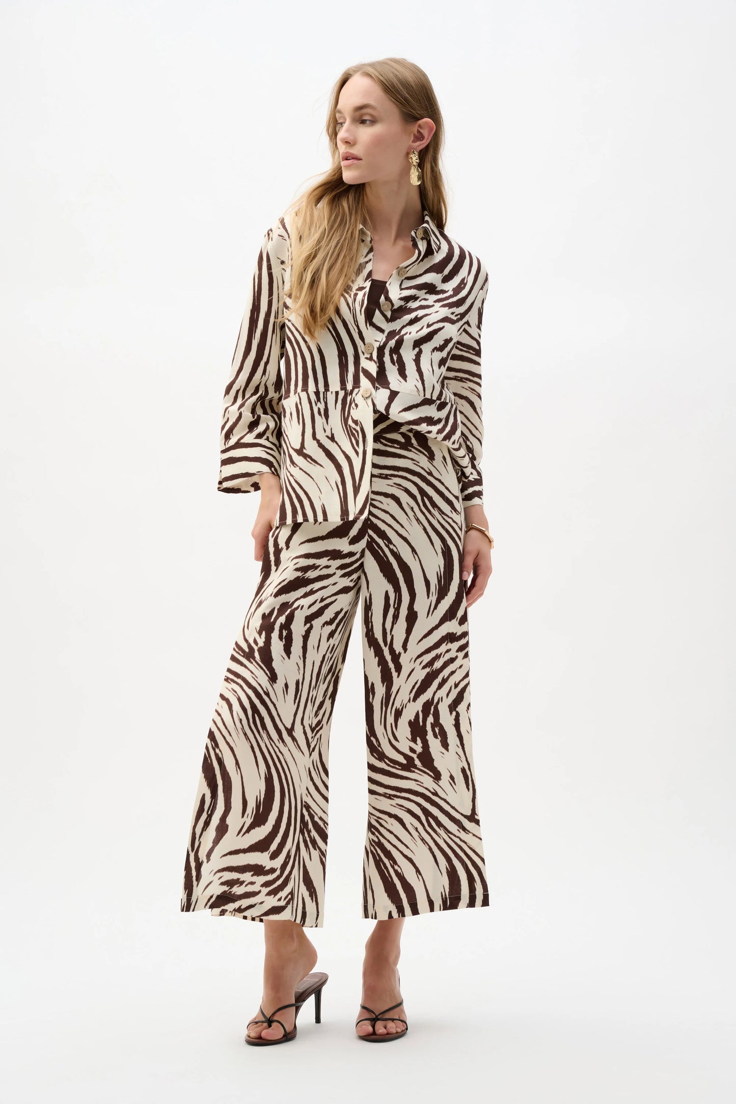 A person stands against a plain background in the Joseph Ribkoff Linen Blend Animal Print A-Line Blouse 262943 and matching wide-leg pants, featuring a bold brown and white animal print for a striking, fashion-forward look.