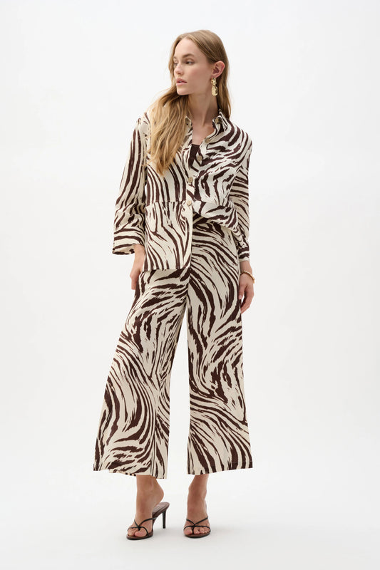 A person stands against a plain background in the Joseph Ribkoff Linen Blend Animal Print A-Line Blouse 262943 and matching wide-leg pants, featuring a bold brown and white animal print for a striking, fashion-forward look.