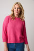 A person with long blonde hair wears the Danbury Top Dahlia Rose 87294 by Parkhurst Cotton Country, made from 100% cotton. They pair it with blue jeans, standing against a plain background, gazing slightly to the side with a neutral expression.