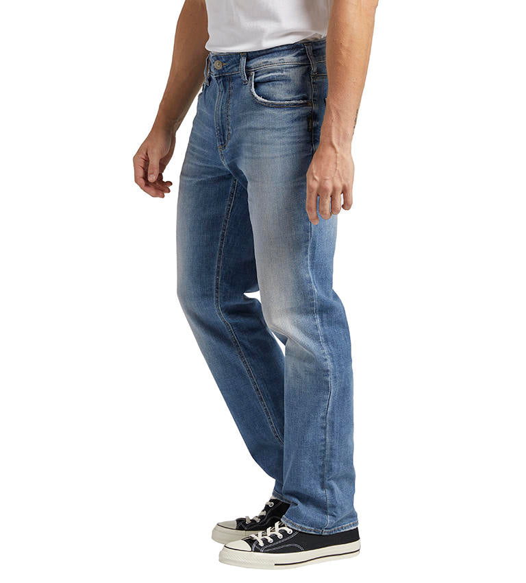 A person is wearing a white shirt tucked into a pair of Grayson Classic Fit Straight Leg Jeans by Silver Jeans Co. These indigo denim men's jeans feature a straight-leg cut with a faded design on the thighs and knees. They are also wearing black and white low-top sneakers.