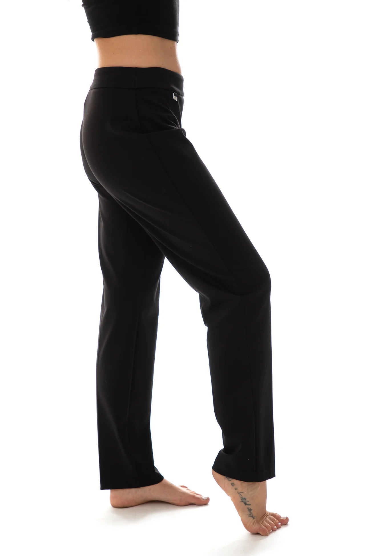 A person is dressed in high-waisted, black slim-fit pants from Bali Corp, specifically the versatile Bali Pant Basic 6451. They are paired with black high-heeled shoes. The background is plain white, keeping the focus on the legs and trousers, while the top part of the person remains out of frame.