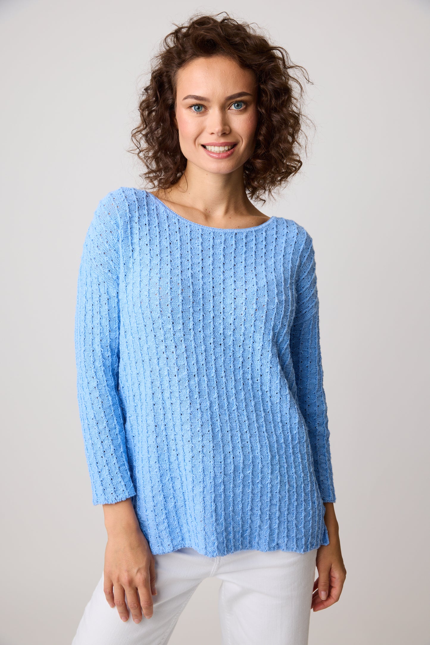 A person with curly hair smiles while wearing the Parkhurst Mia Tunic Very Peri, and white pants. They are standing against a plain, light-colored background.