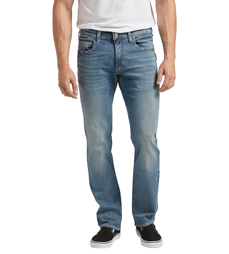 A person is standing sideways, dressed in Allan Slim Fit Straight Leg Jeans from Silver Jeans Co., a white T-shirt, and black slip-on shoes. The jeans have a fitted silhouette with slight fading on the thighs and knees. The person's arm is relaxed by their side.