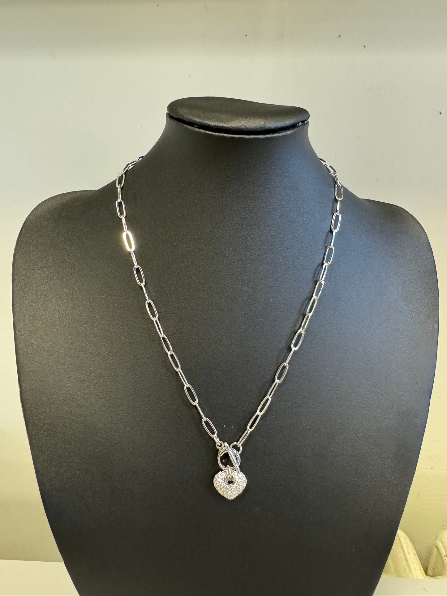 The Fiona 23 Necklace from Elise Accessories, featuring a silver chain with elongated links and a glittering heart-shaped pendant, is elegantly showcased on a black mannequin bust against a simple backdrop. This remarkable piece embodies the timeless elegance that defines fashion.