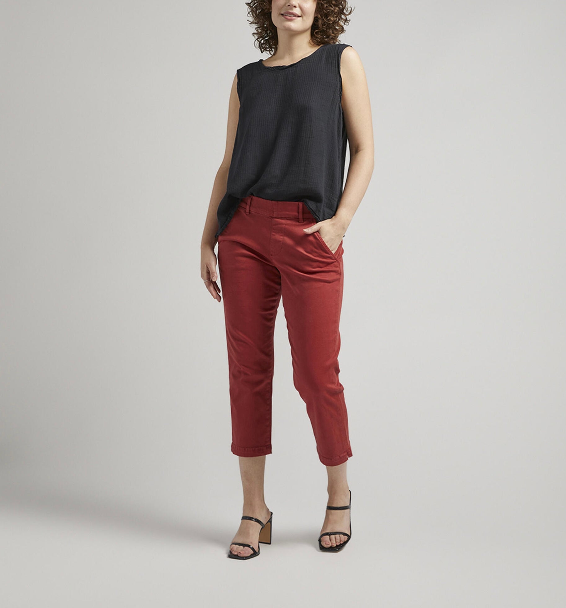 A person stands against a plain backdrop with a neutral expression. They are wearing a sleeveless black top, flattering Maddie Capri Mid Rise pants in brick-red from JAG, and black heeled sandals. Their left hand rests in their pocket, and they have curly hair.