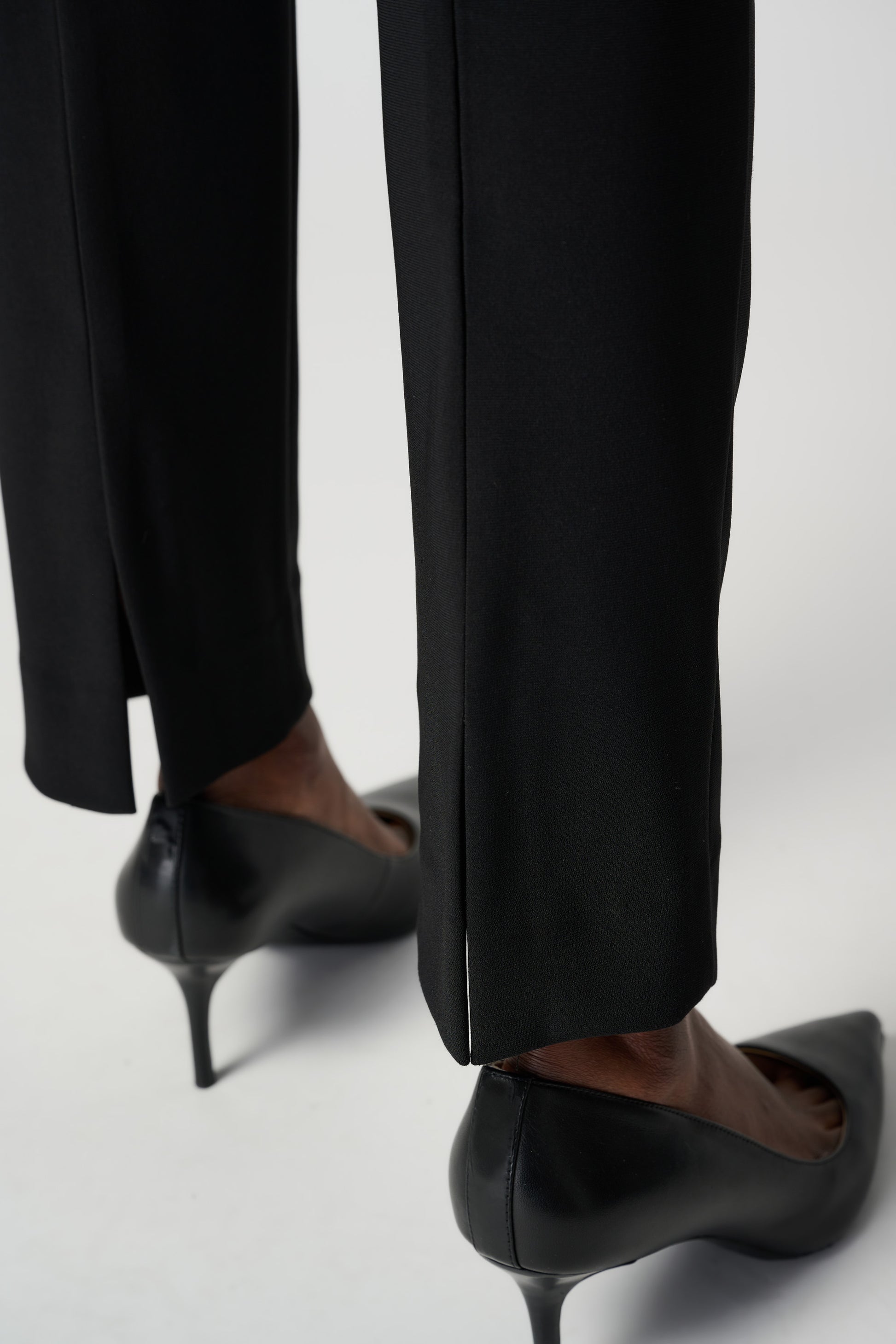 A person wearing the Classic Straight Pant in black by Joseph Ribkoff with slits at the ankles and black pointed heels, standing with their back to the camera. The person is also wearing a white top made of silky knit material. The background is plain white.