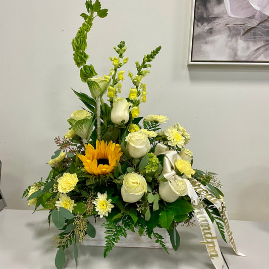 The "Warm Remembrance" funeral arrangement by Posies Flowers & Fashion features white roses, yellow blooms, a vibrant sunflower, and lush green foliage arranged in a white vase. A ribbon with "Grandpa" written on it adorns the bouquet. The arrangement is displayed on a gray surface against a light-colored wall next to a wooden box.