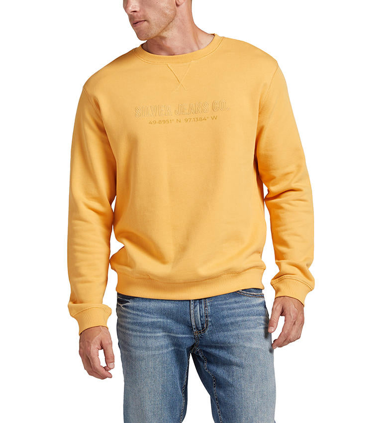 A man wearing a "HONEY" French terry cotton men's crewneck sweatshirt by Silver Jeans Co., featuring "49.8951 N -97.1384 W" embroidered on the front, paired with blue jeans. The sweatshirt includes ribbed cuffs and a crew neckline. He is looking slightly to the side, with one hand by his side and the other partially in a pocket.