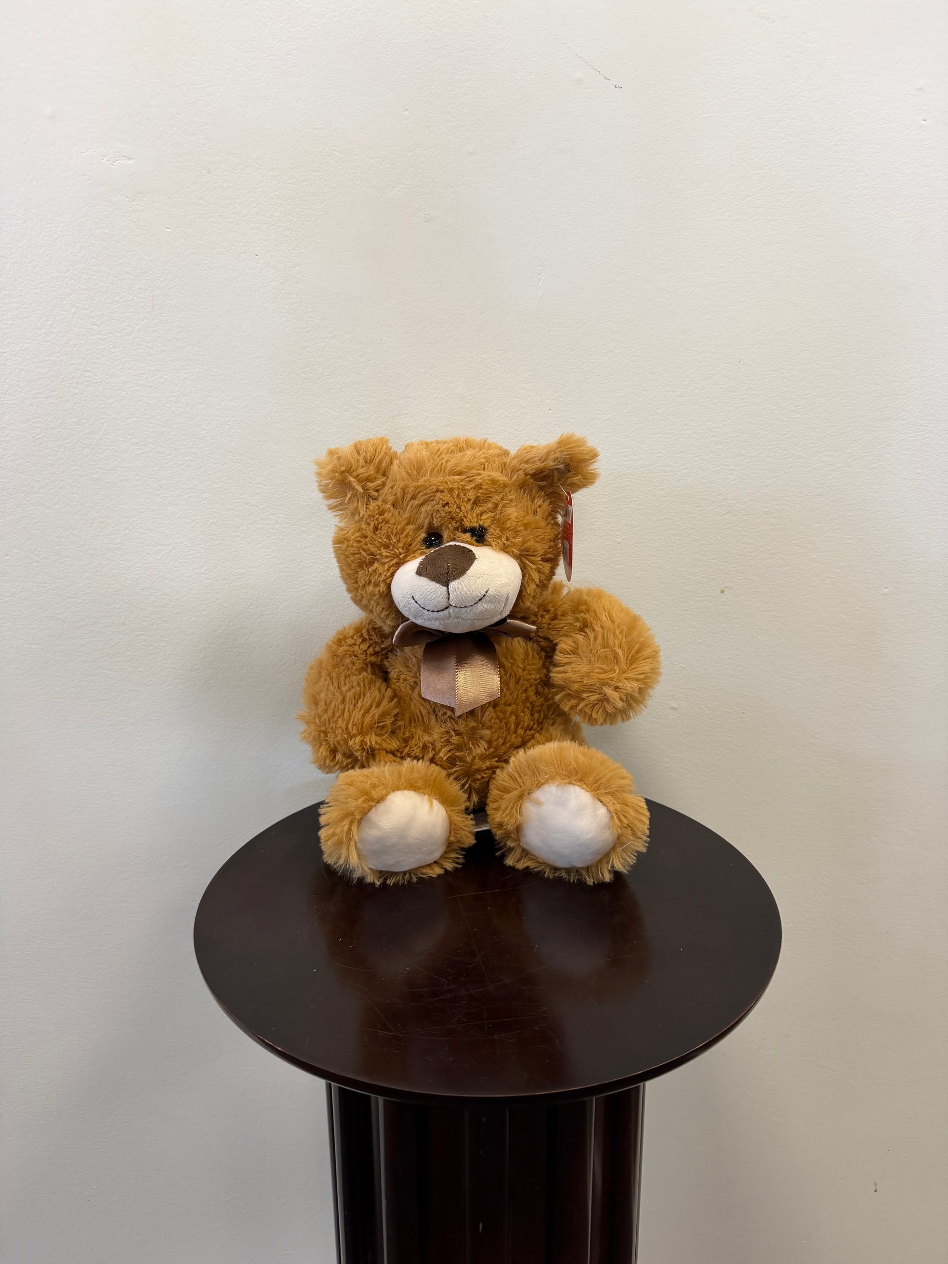 A Classic Teddy Bear – Fiesta by Fiesta, featuring a beige snout and a bow, sits on a dark round pedestal against a plain light-colored wall—making it a charming choice for gift delivery.