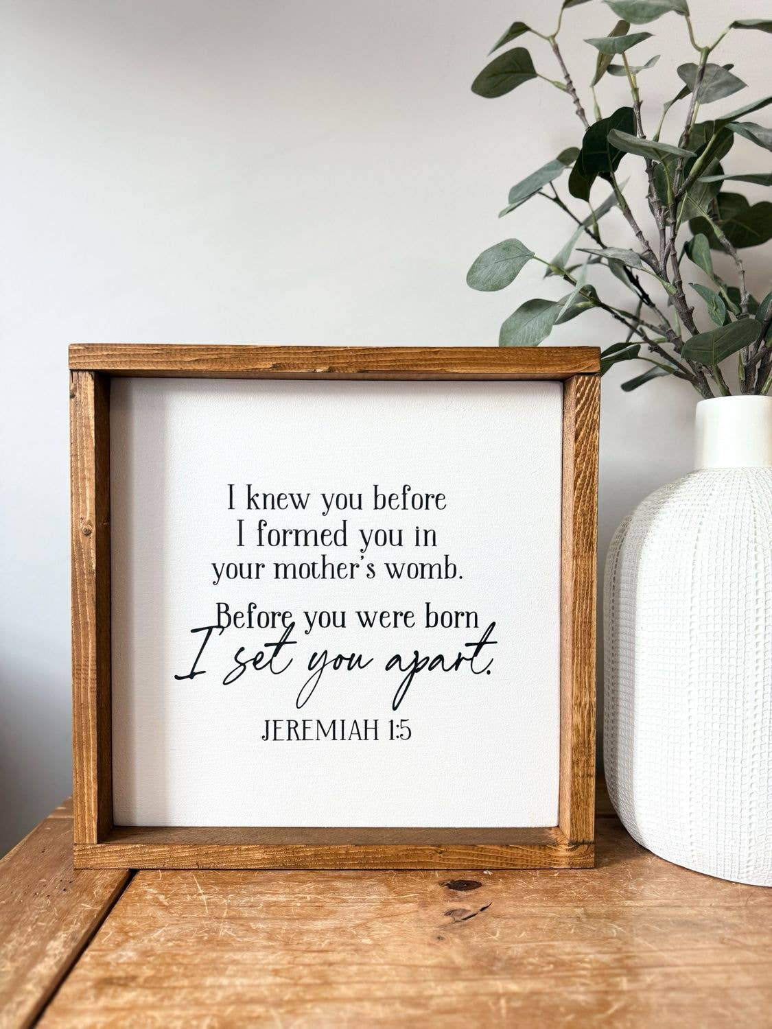 An “I Knew You Before… I Set You Apart” Wood Sign by Inspired Findings sits on a wooden table beside a white textured vase filled with green leafy branches. This elegant wood framed sign from Inspired Findings offers unique home decor, serving as an inspiring focal point against the plain white background.