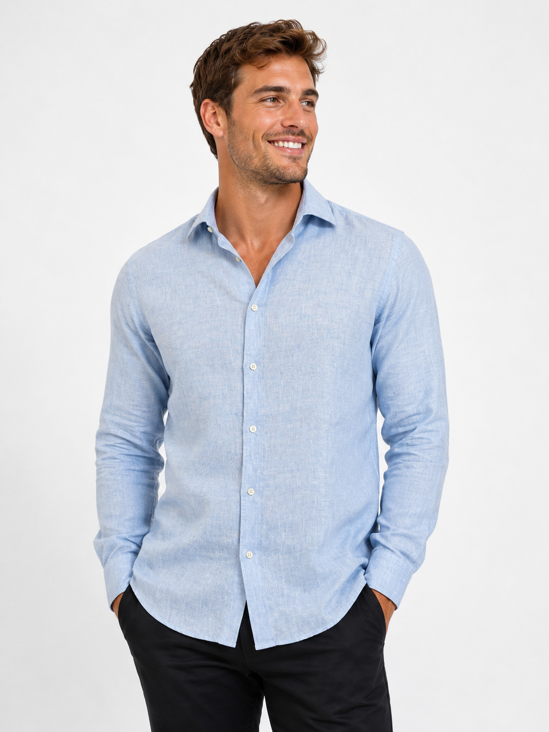 A man wears the Paul Miranda Linen Shirt Cielo and black pants, standing against a white background, smiling and looking left with his hands in his pockets.