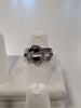 A shimmering Jenna-57 Adjustable Ring by Elise Accessories, showcasing a sophisticated silver finish with an oval-shaped, encrusted band design on top, is displayed vertically on a white ring stand. The background is blurred, accentuating the intricate details and sparkle of the elegant CZ-adorned ring.