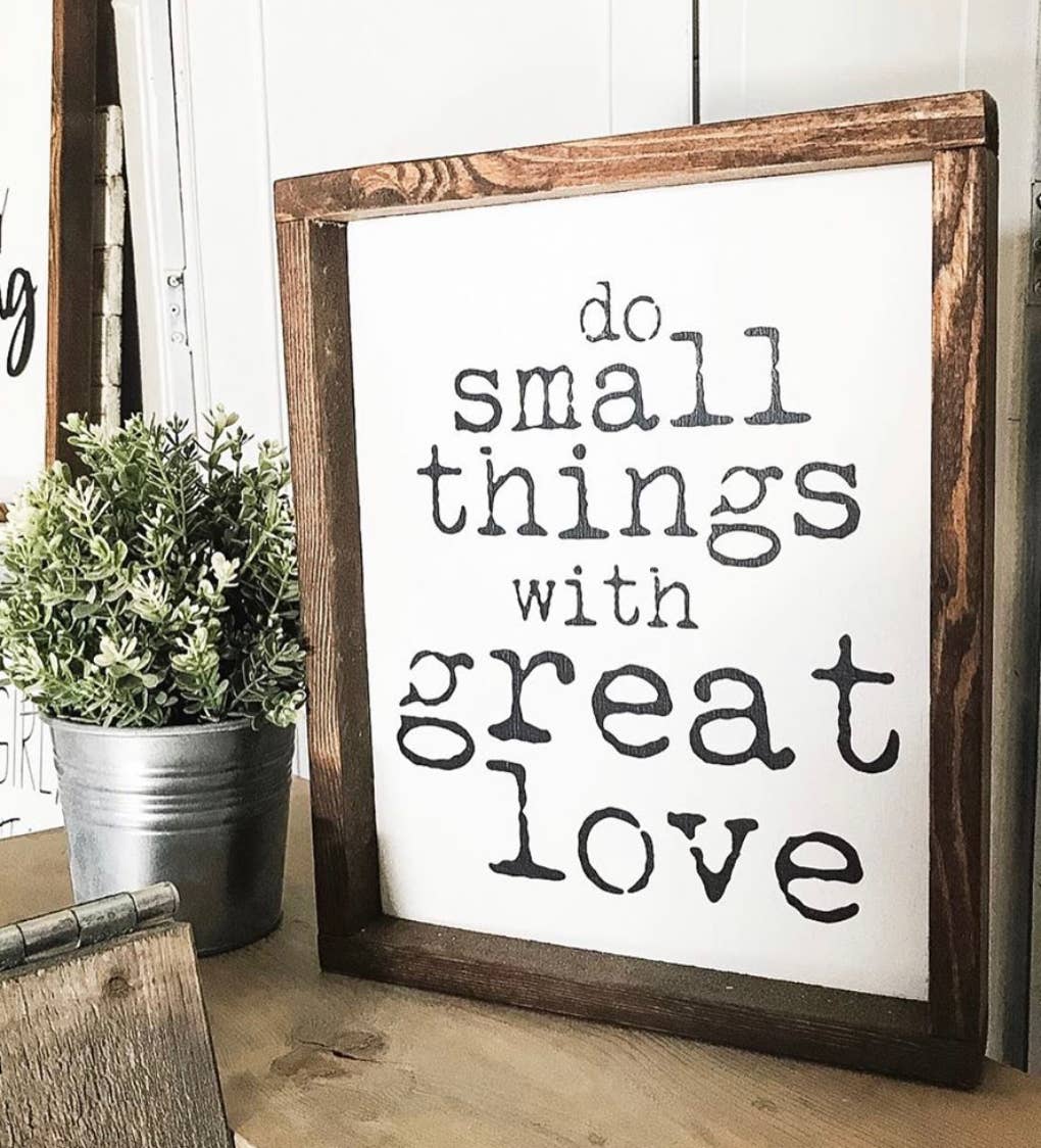 A Do Small Things Wood Sign Sm by Inspired Findings rests beside a potted plant on a wooden surface, infusing the space with rustic charm and an uplifting message.
