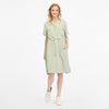 A woman in a light green ROISANA DRESS 2511-20026-5036 by RAGWEAR smiles with one hand in her pocket. The dress, made from 100% viscose, has a drawstring waist and short sleeves. She pairs it with white sneakers, and her wavy blonde hair stands out against the plain white background.