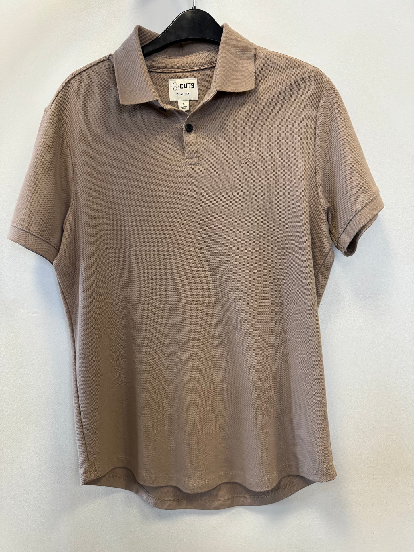 A light brown Universal COZ Polo - CURVE HEM from Once We Were Warriors hangs on a black hanger against a plain white wall. This short-sleeved Pima cotton shirt features a collar, a three-button placket, and a subtle embroidered logo on the left chest. With its modern, casual style and soft pique texture, it's perfect for everyday wear.