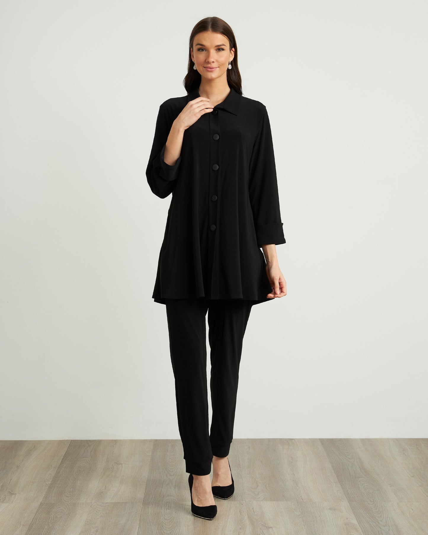 An individual wearing Joseph Ribkoff's Drawstring Waist Pants Style 211317, featuring zipper detailing in black, paired with a white top and black heels. The person stands on a wooden floor against a plain, light-colored background, seamlessly combining comfort and style.