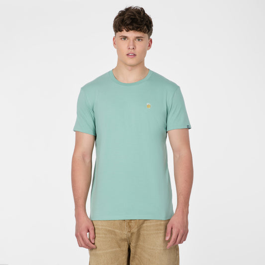 A young man with short brown hair wears the RAGWEAR T-shirt NEDDIS C 2612-15023-5036 in Dusty Green, paired with tan shorts, standing against a white background and embodying vegan streetwear style.