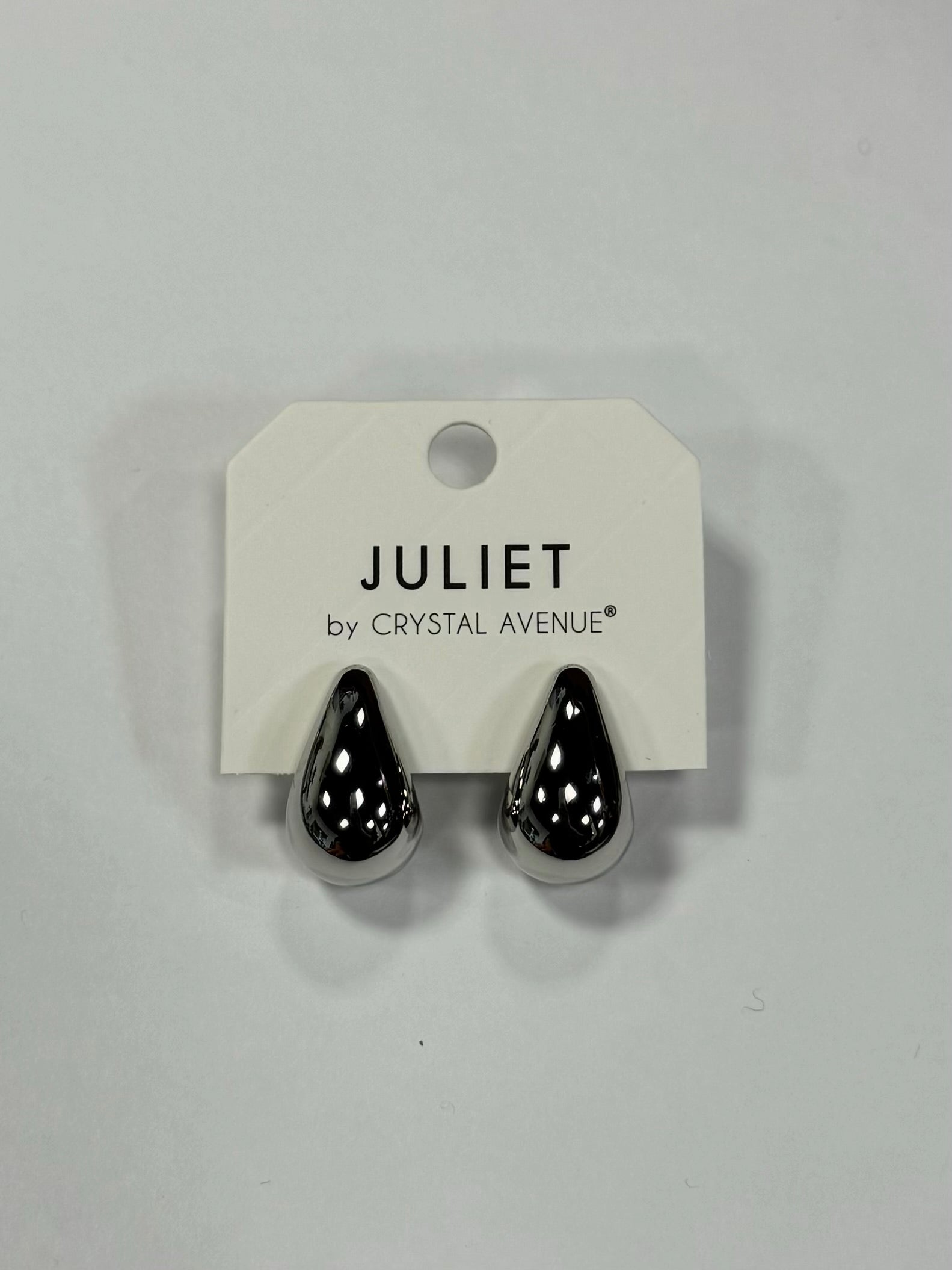 A pair of stylish, shiny, silver-tone teardrop earrings is displayed on a white tag labeled "Elise Accessories by Posies Flowers & Fashion Inc.," evoking the elegance of Dior Earrings against a plain background.