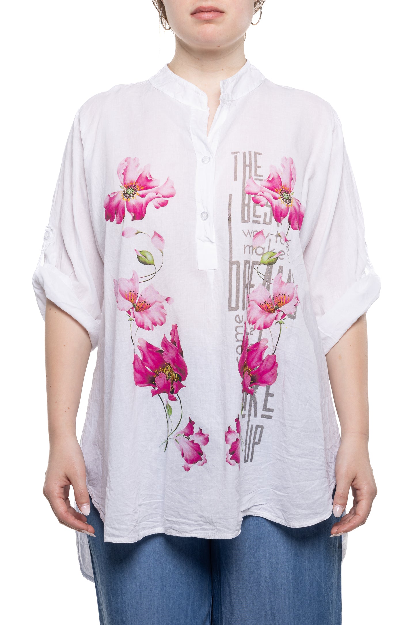 A person wearing an ETERNELLE BUTTON COTTON SHIRT FLORAL stands against a white background. The loose, 100% cotton shirt, adorned with bright pink floral designs and text that reads "THE BEST way to DREAM" among other phrases, has long sleeves and is paired with blue jeans.