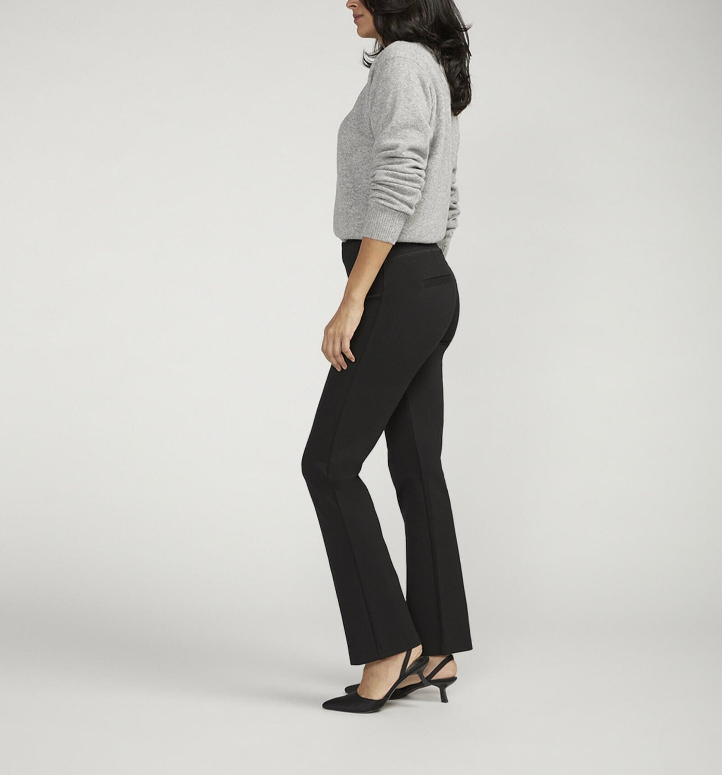 A person is posing against a plain background, wearing a light gray long-sleeve sweater paired with JAG's Mid Rise Bootcut Pull-On Pants. They have one hand on their hip and are sporting black pointed-toe heeled shoes. Their dark hair flows loose and wavy.