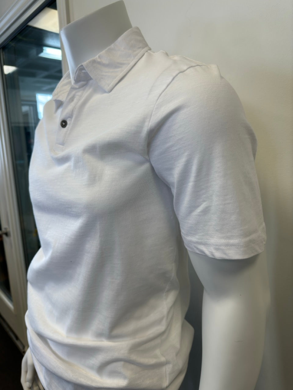 A mannequin wearing the Cotton Polo Men’s in white, featuring a classic collar and short sleeves, with three dark buttons on the placket, stands against a plain, light-colored background. The outfit is completed with dark pants from Silver Jeans Co., showcasing a casual yet stylish look.