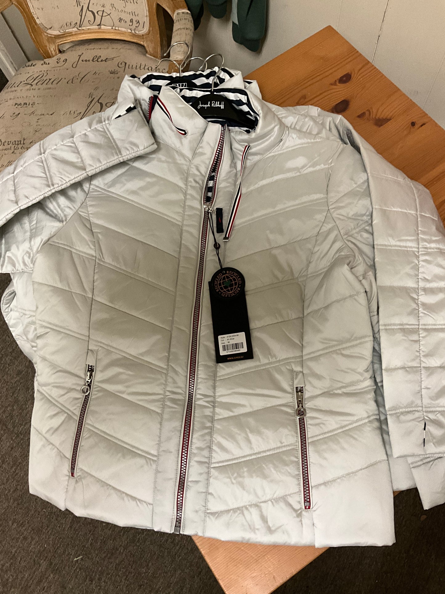 The JUNGE 91-Silver Puff Jacket 0126-2254-66, a light gray quilted jacket with red and white trim on the zippers and collar, is displayed flat on a wooden surface with a black tag attached to the front zipper.