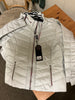 The JUNGE 91-Silver Puff Jacket 0126-2254-66, a light gray quilted jacket with red and white trim on the zippers and collar, is displayed flat on a wooden surface with a black tag attached to the front zipper.