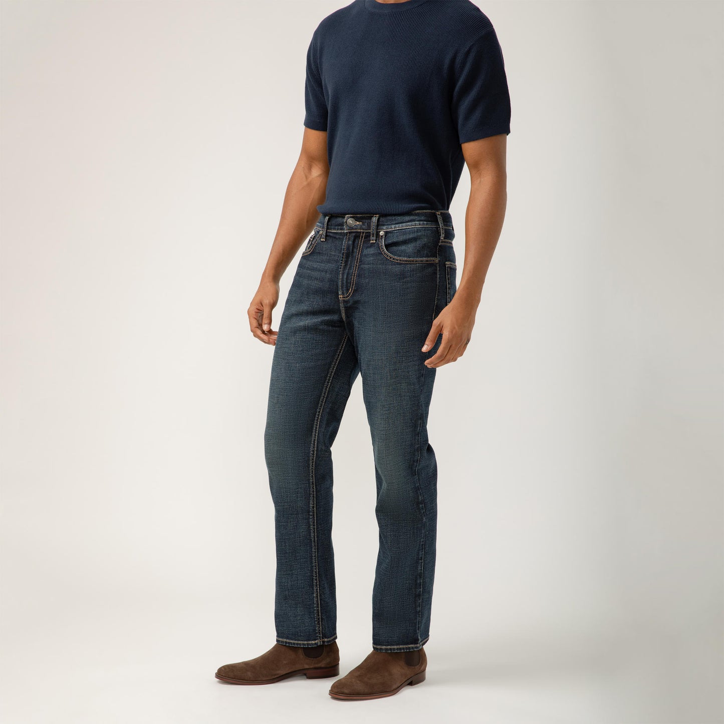 A person stands against a plain background wearing a navy blue, classic fit, short-sleeve shirt tucked into Grayson Classic Fit Straight Leg Silver Jeans M23425EFS409 by Silver Jeans Co. with brown suede shoes. Their arms rest comfortably by their sides, and their shoes contrast with the white backdrop. Style code: SIL-M23425EFS409-IND.