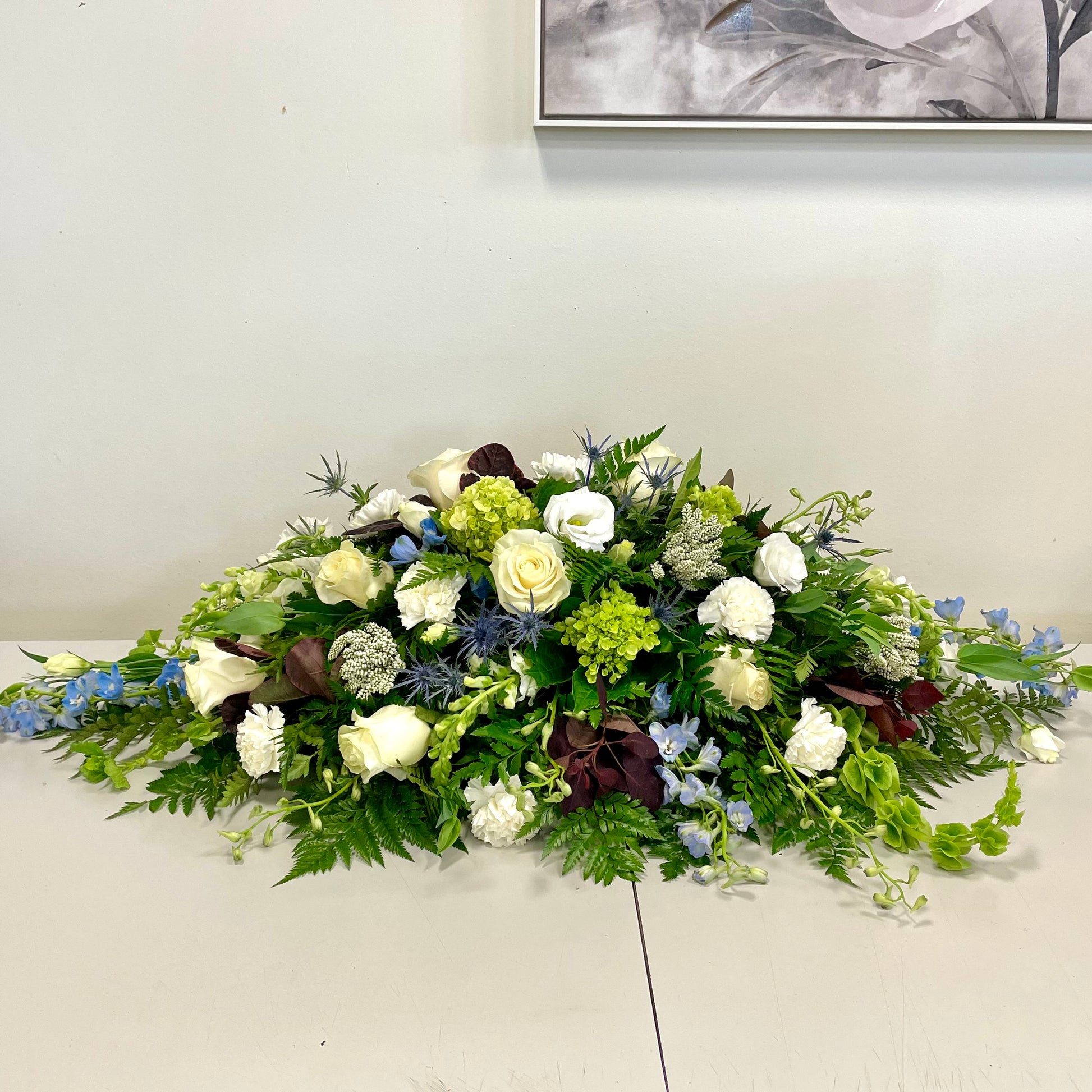 The "Tranquil Embrace" funeral casket spray by Posies Flowers & Fashion, featuring a beautiful mix of white roses, hydrangeas, blue delphiniums, and various greenery, is arranged horizontally on a white surface against a light-colored wall. Part of a framed artwork is visible in the background.