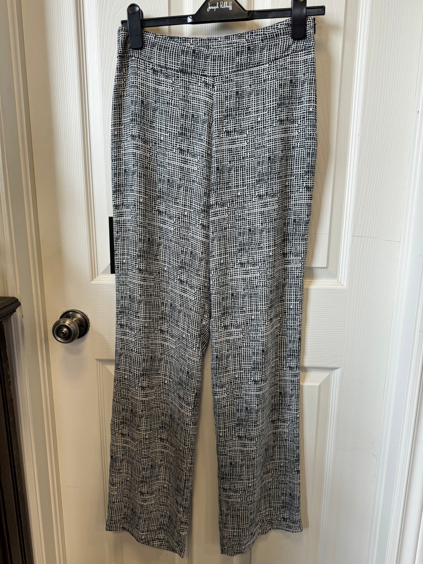 A pair of white and black plaid dress pants hangs on a black plastic hanger against a white door with a silver doorknob. The "DK" logo on the hanger hints at these Tribal Paz Torras - Talla Dress Pants V17410-4 adding to your sophisticated wardrobe.