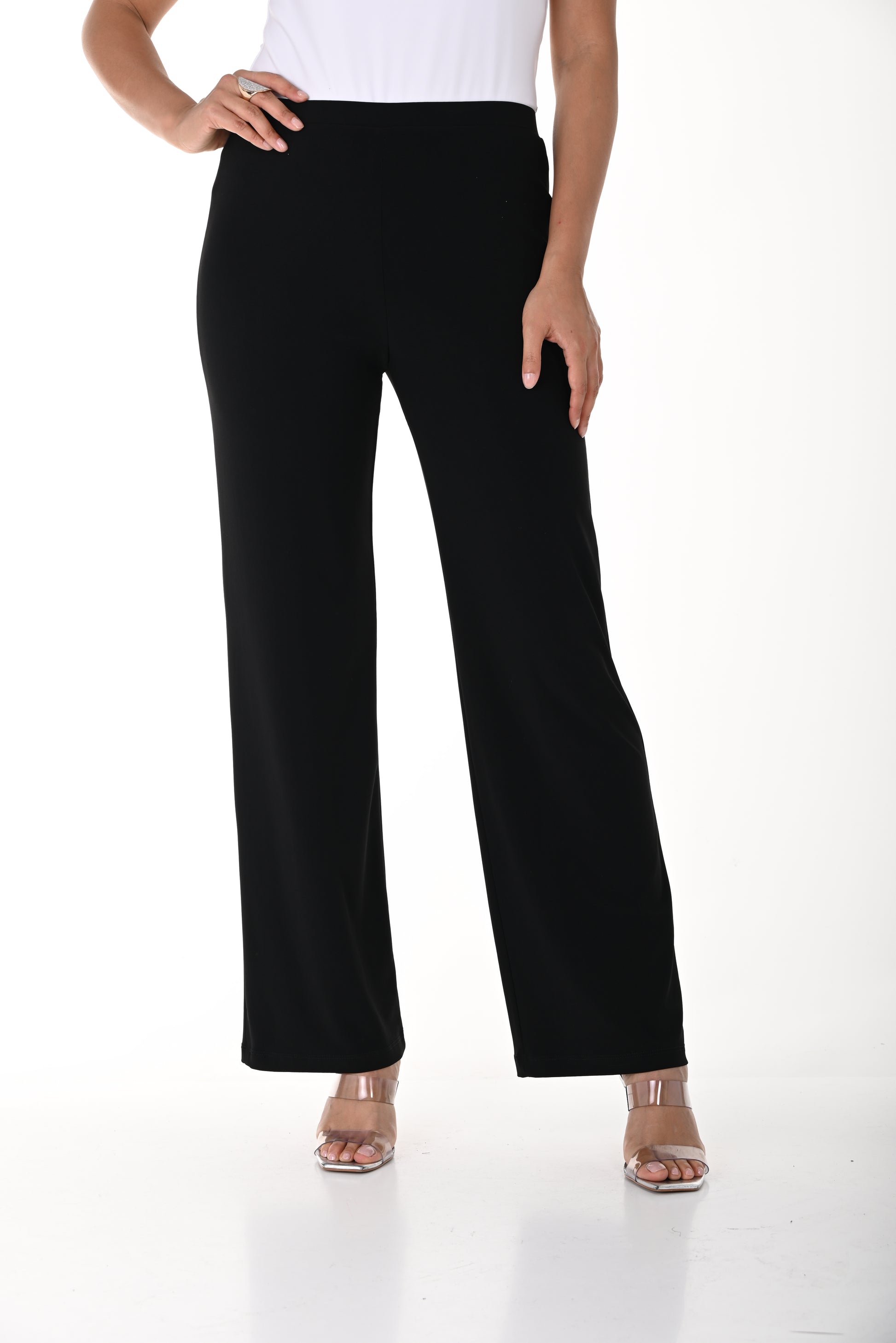 A person models the Frank Lyman Classic Pull-On Pant – Black (Style 006), featuring a straight-leg silhouette, paired with open-toed heels and standing against a white background. Their upper body is partially visible, with one hand resting on their hip.