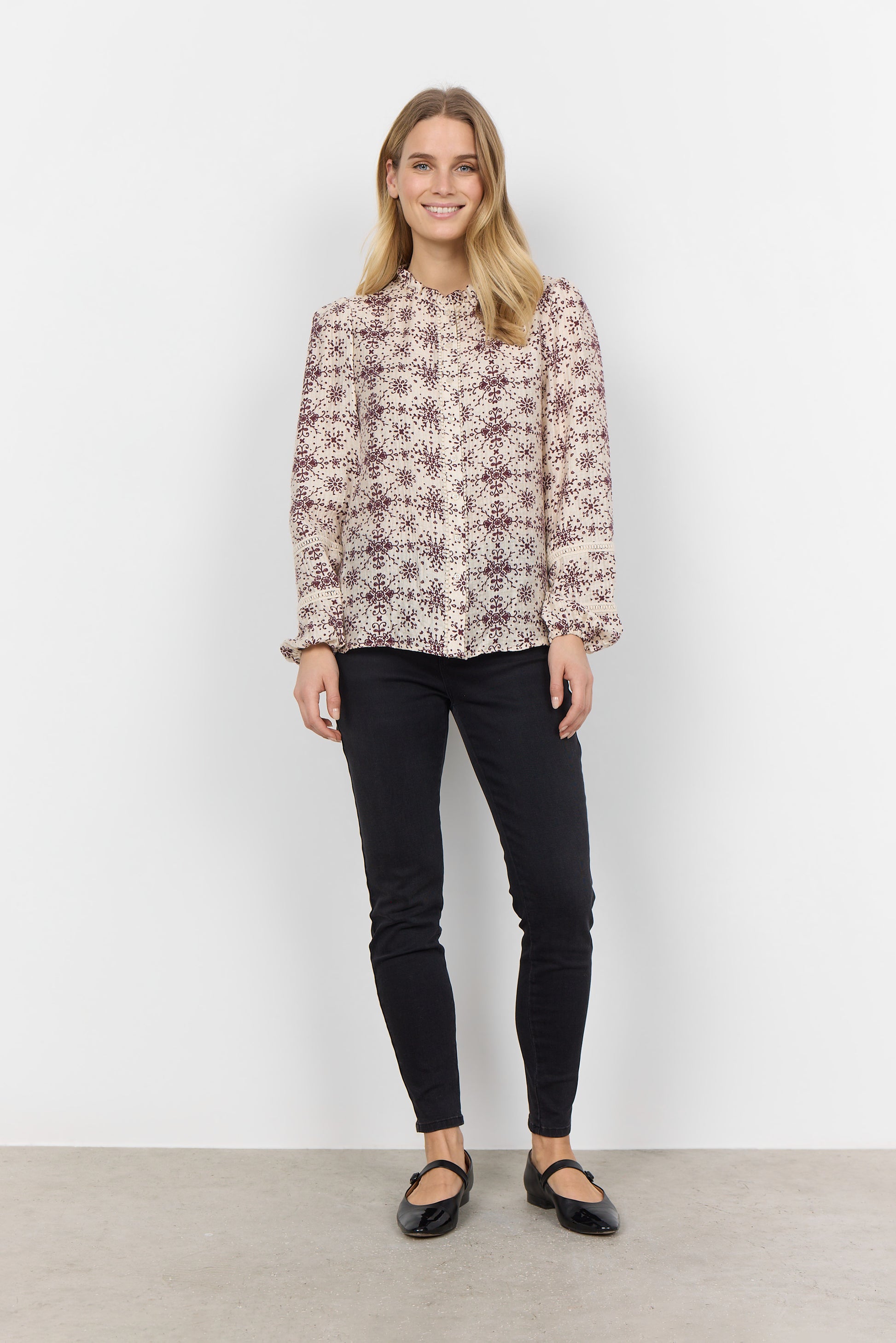 A woman with long blonde hair is wearing the luxurious blend of fabrics in the Shirt SC-IANE 2 from RAGWEAR, featuring a cream-colored blouse with a maroon floral pattern and black pants. She stands against a white background, looking directly at the camera with a neutral expression.