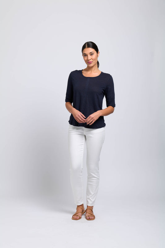 A woman stands against a plain light background, wearing the FOIL Heroine Top CFO7824 -FOIL in navy blue with white pants and tan sandals. Her hair is tied back and her hands are gently clasped—perfect for a stylish summer look.