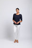 A woman stands against a plain light background, wearing the FOIL Heroine Top CFO7824 -FOIL in navy blue with white pants and tan sandals. Her hair is tied back and her hands are gently clasped—perfect for a stylish summer look.