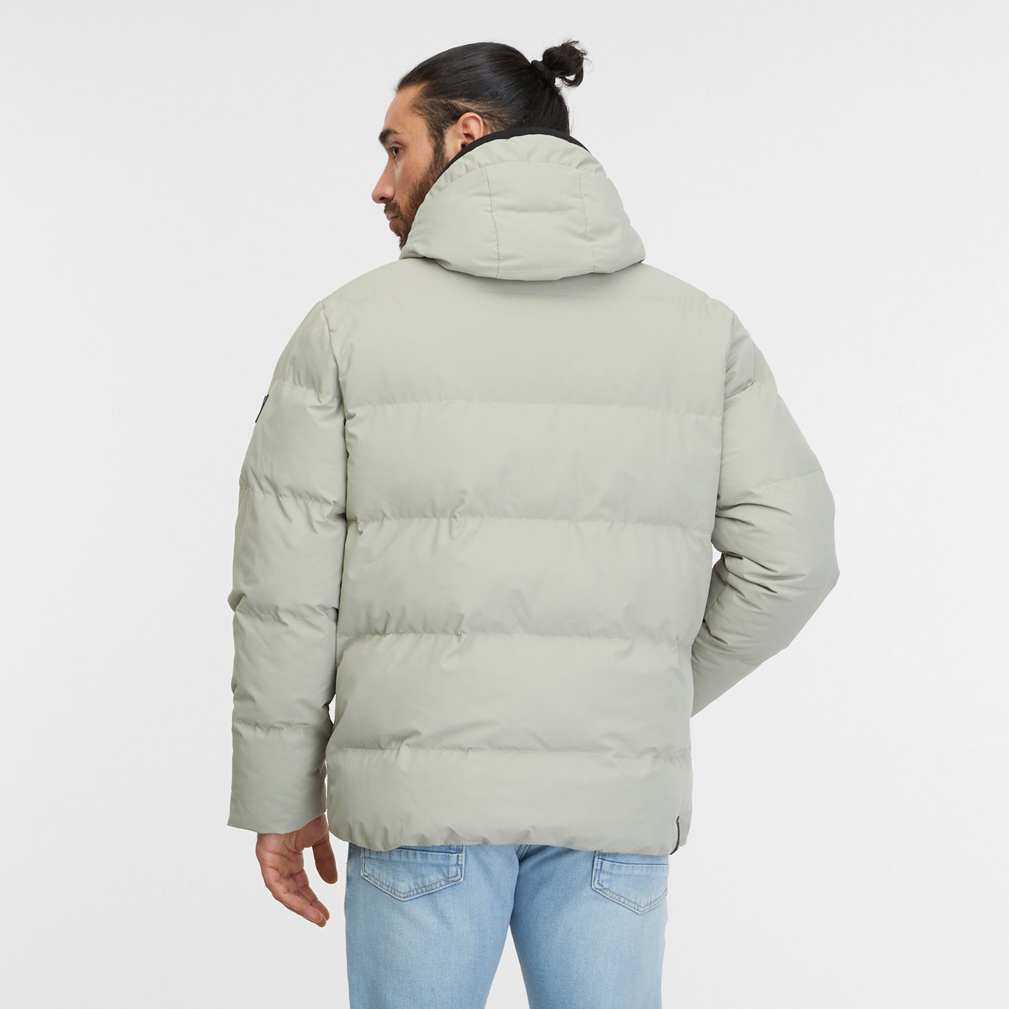 A man with long hair tied back in a bun is wearing a dusty olive Puffer Coat North from RAGWEAR and light blue jeans. He is standing against a plain white background, with his right hand in his jacket pocket and a serious expression on his face, epitomizing vegan streetwear.