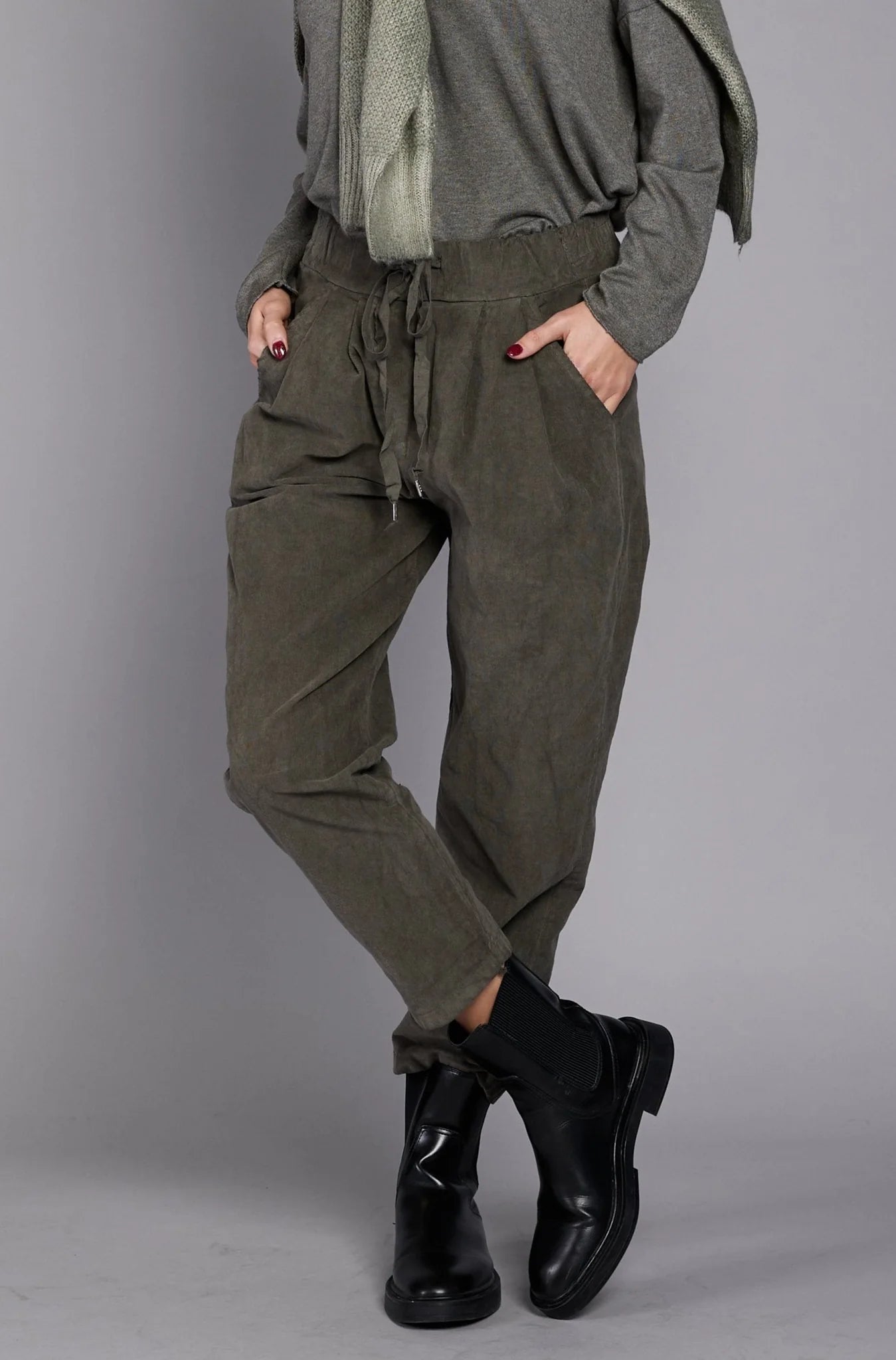 A person wearing a gray long-sleeve top, the stylish EL27901 pants by ELISSIA in ankle-high olive green with a drawstring, and black ankle boots stands with their hands in their pockets against a plain grey background. A scarf is draped over their shoulder. Only the lower half of the person is visible.