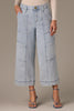 Wearing Skyrise Double Bottom Wide Leg Jeans by Wit & Wisdom, a person stands with hands in pockets. The light denim, high-waisted jeans feature large pockets and are paired with a beige long-sleeve top and blue strappy sandals against a plain grayish background.