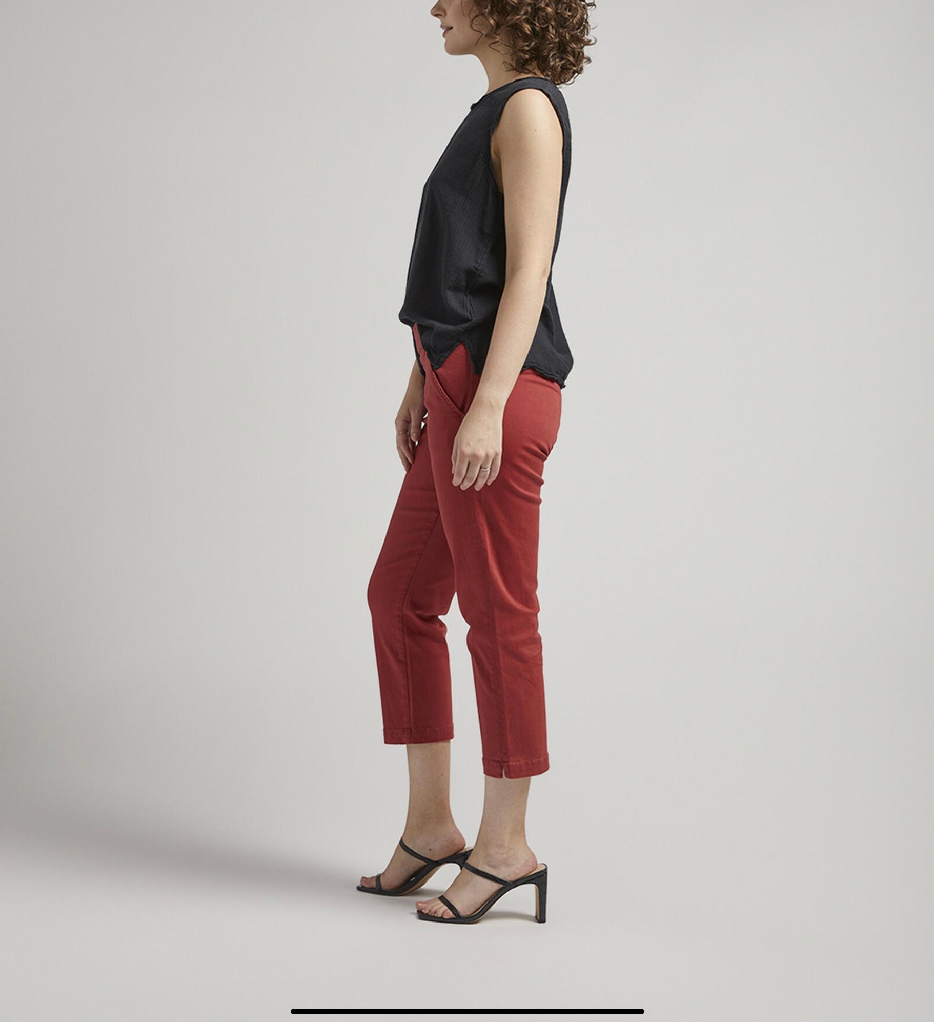 A person stands against a plain backdrop with a neutral expression. They are wearing a sleeveless black top, flattering Maddie Capri Mid Rise pants in brick-red from JAG, and black heeled sandals. Their left hand rests in their pocket, and they have curly hair.