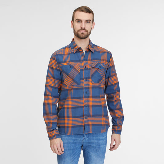 A man with a beard is standing against a plain white background. He is sporting a blue and orange plaid "Shirt Checki Blue 2422-40003-2040" by RAGWEAR, a vegan streetwear cotton flannel shirt with two chest pockets, paired with blue jeans. He is looking straight at the camera with a neutral expression.