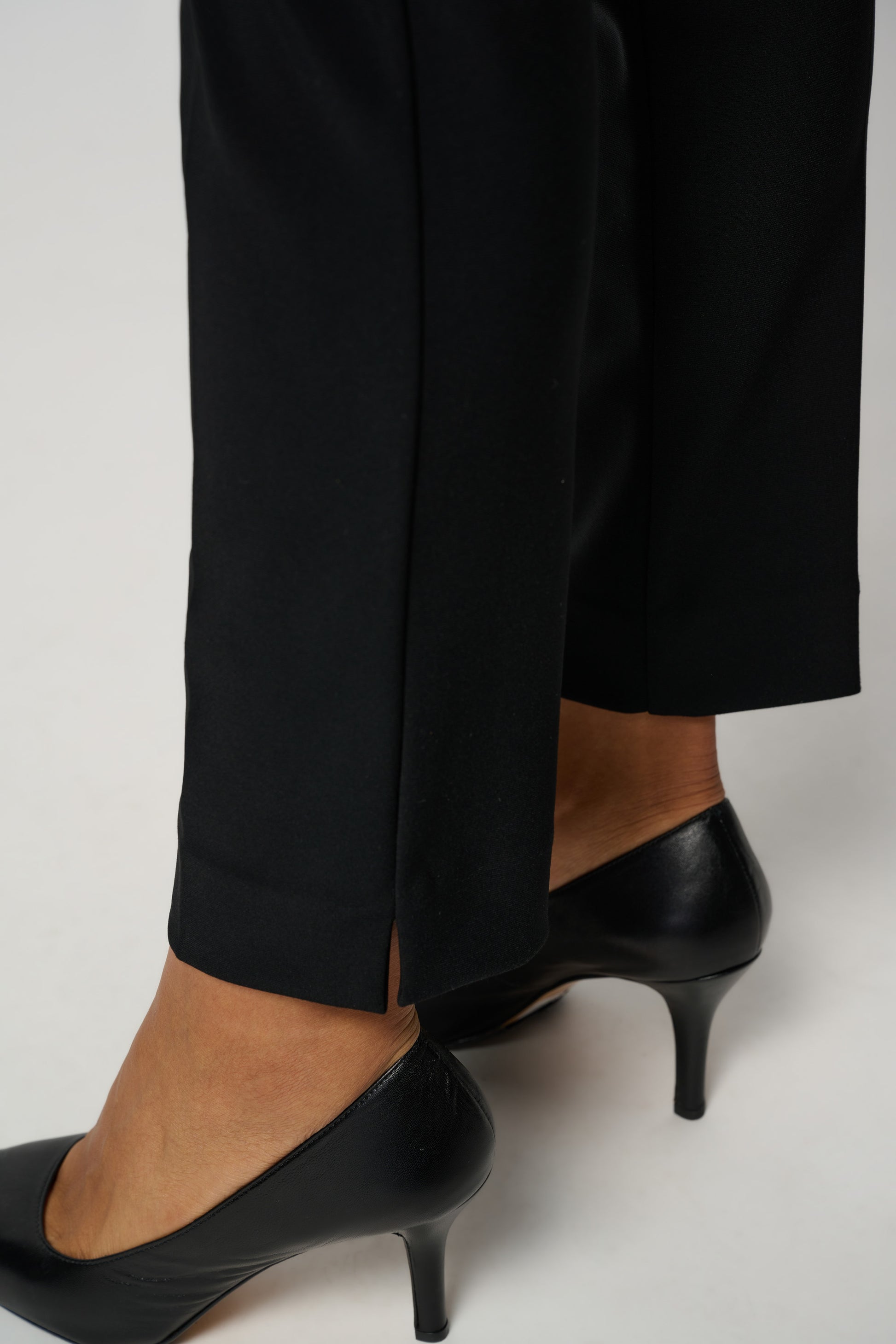 A person is shown from the side wearing Joseph Ribkoff's Classic Tailored Slim Pant, adorned with a JR tab ornament, paired with black high-heeled shoes. The background is plain white, and the person is also wearing a white top that is partially visible in the image.